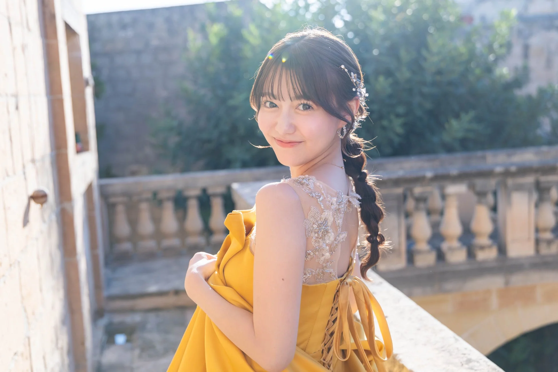 黄色の華やかなドレスを身につけ、編み込みヘアに髪飾りをつけた女性が、振り返って笑顔を見せているポートレート。美しい逆光の中で、上品な雰囲気が漂う。