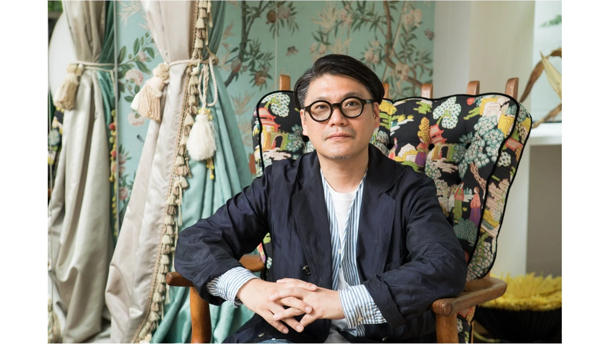 眼鏡をかけた男性が、華やかな柄の椅子に座りカメラを見つめているポートレートの画像