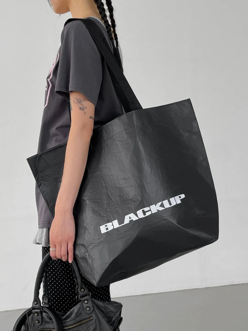 グレーのTシャツとドット柄のボトムスを着用した女性が、肩に大きな黒いトートバッグ（BLACKUPのロゴ入り）と手に小さな黒いレザーバッグを持っています。