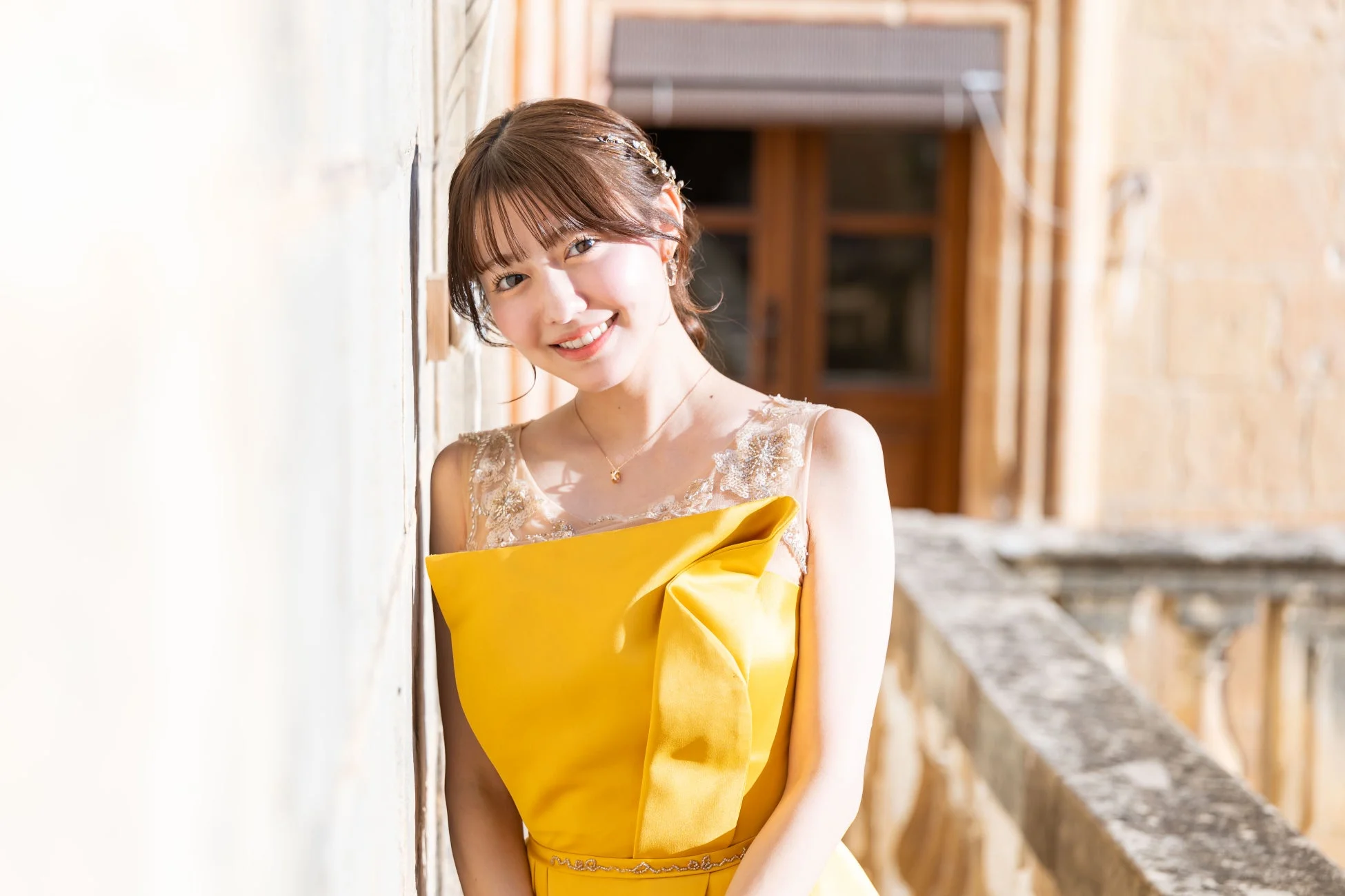 黄色のエレガントなドレスを着た若い女性が、屋外の壁にもたれかかって優しく微笑んでいます。ヘアアクセサリーとネックレスが華やかさを添え、明るい日差しの中で美しいポートレートとなっています。