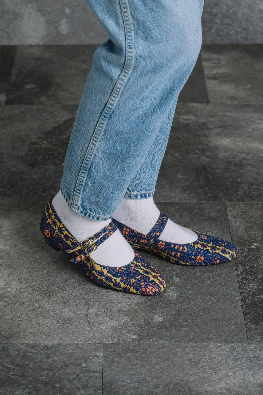 明るい色のジーンズと白い靴下を着用した人物の足元が写っています。特徴的なのは、青と黄色の複雑な模様があしらわれたメリージェーンスタイルのフラットシューズです。背景は暗い色のタイル張りの床です。