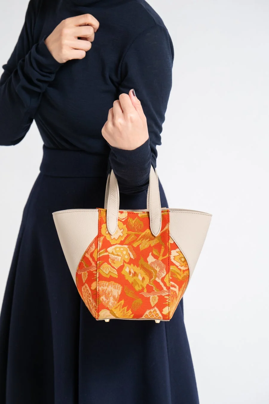 紺色のトップスとスカートを着用した女性が、鮮やかなオレンジ色の花柄とベージュの組み合わせが特徴的なハンドバッグを持っている様子。シンプルながらもデザイン性のあるバッグが際立つファッション写真です。