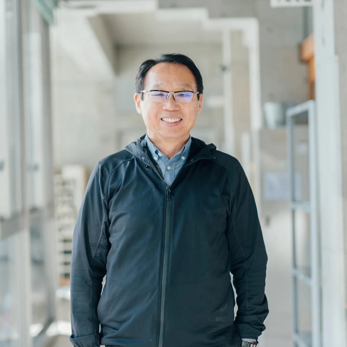 眼鏡をかけたアジア系の男性が、黒いジャケットと青いシャツを着て、明るい屋内で笑顔で立っているポートレート写真です。親しみやすい表情が印象的です。