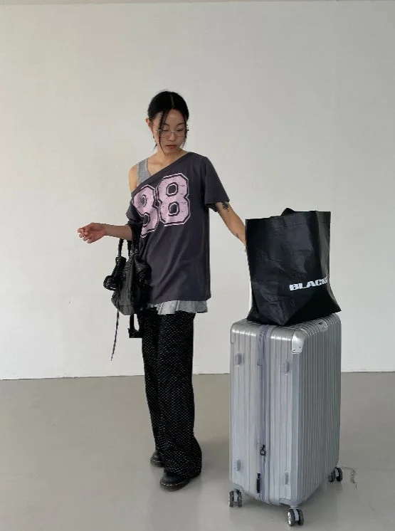 白い壁を背景に、肩出しTシャツとワイドパンツを着た女性がスーツケースの横に立っています。