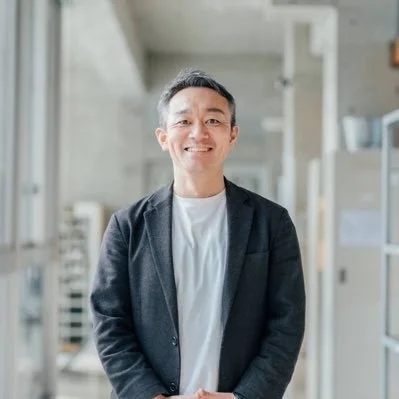 明るい表情でカメラを見つめる中高年のアジア人男性のポートレートです。ダークなジャケットと白いTシャツを着用し、背景はオフィスや廊下のような屋内空間がぼやけて写っています。