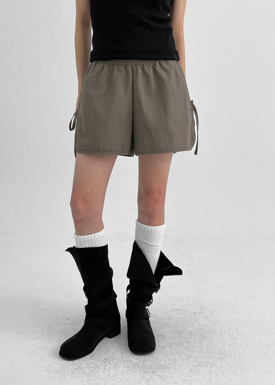 白い背景の前で、黒いノースリーブトップスにオリーブグリーンのショートパンツ、白いソックスと黒いミドル丈ブーツを合わせた女性のファッションスナップです。