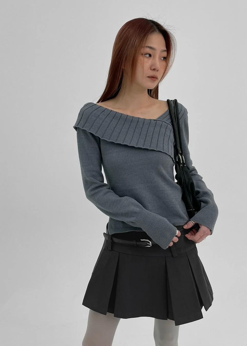 白い背景の前でポーズをとる若い女性。グレーのオフショルダーニットとダークグレーのプリーツミニスカートを着用し、黒いショルダーバッグを肩にかけています。