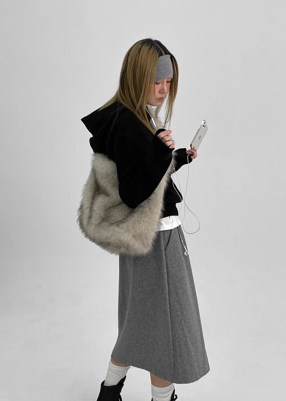 黒いパーカーとグレーのスカートを身につけ、ファーバッグを肩にかけた女性が、イヤホンで音楽を聴きながらスマートフォンを見ています。